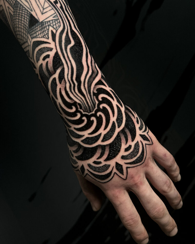 Tatuaje blackout de líneas onduladas y pétalos geométricos entre el antebrazo y la mano, en negro